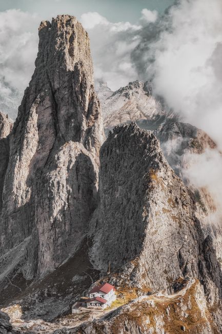 Breathtaking view of the Dolomites with rugged peaks and an alpine hut nestled below, enveloped by clouds.