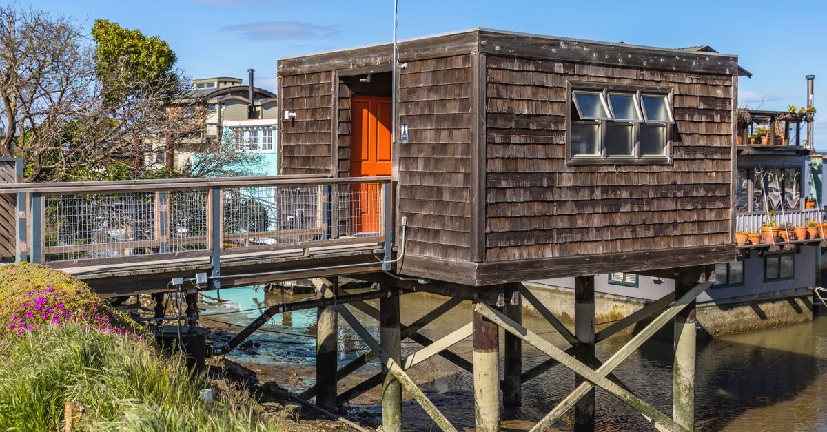 A charming wooden stilt house with a vibrant orange door, nestled by a tranquil river under a clear blue sky.