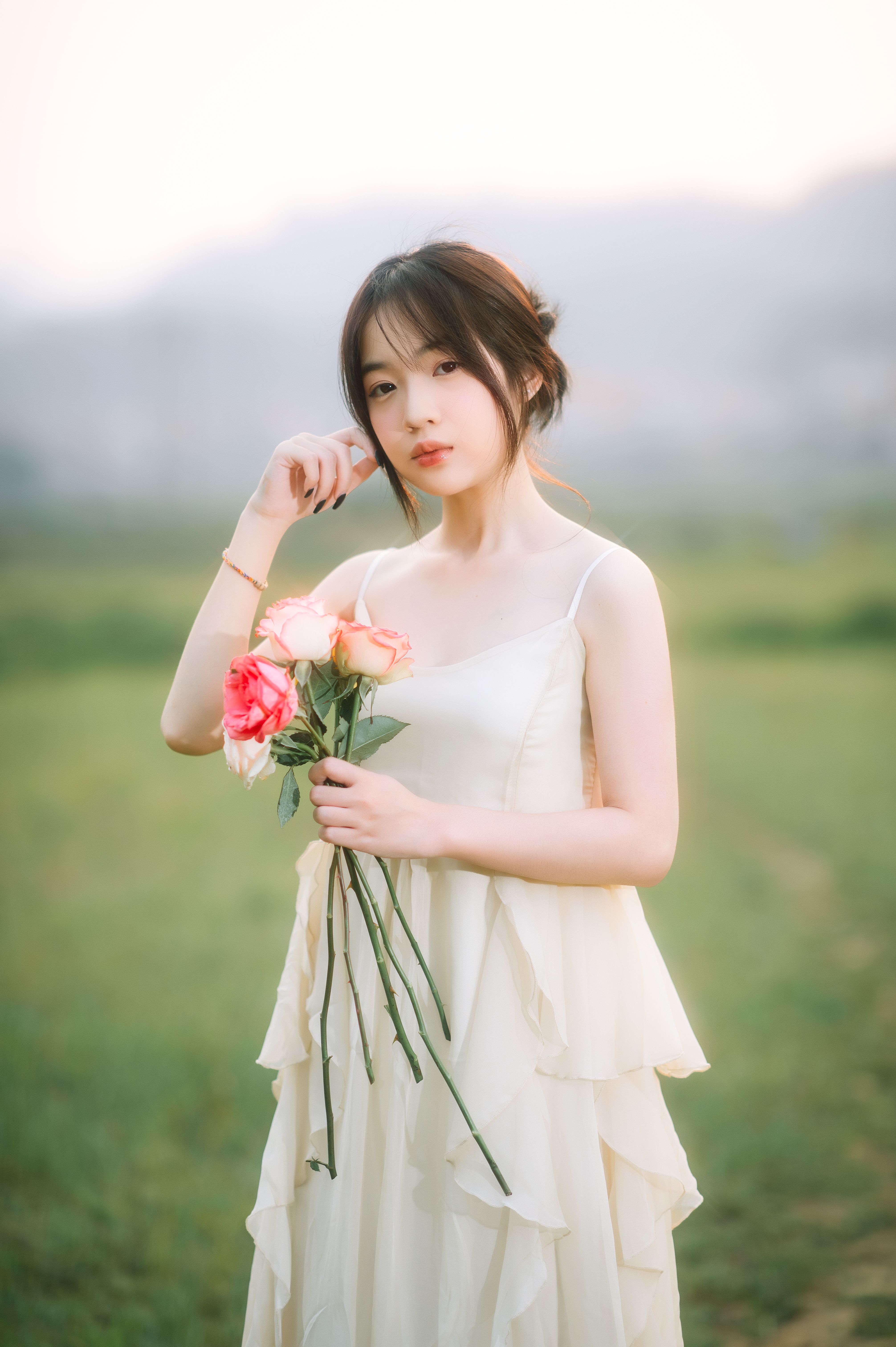 A young woman in a white dress holding roses stands gracefully in a lush green field, exuding elegance and tranquility.