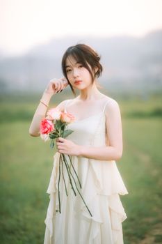 一位穿着白色裙子的年轻女性手持玫瑰，优雅地站在郁郁葱葱的田野中，散发着优雅和宁静。 - 相关推荐