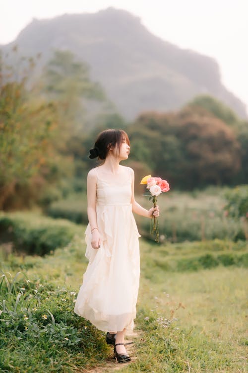 Young Woman in a White Dress Walking on a Field and Holding Flowers