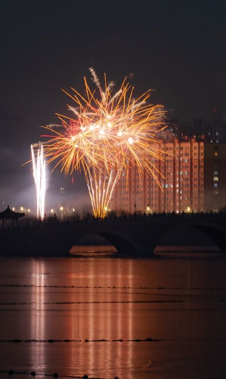 Free Vibrant fireworks illuminate the night sky over a city bridge, reflecting on the river below. Stock Photo