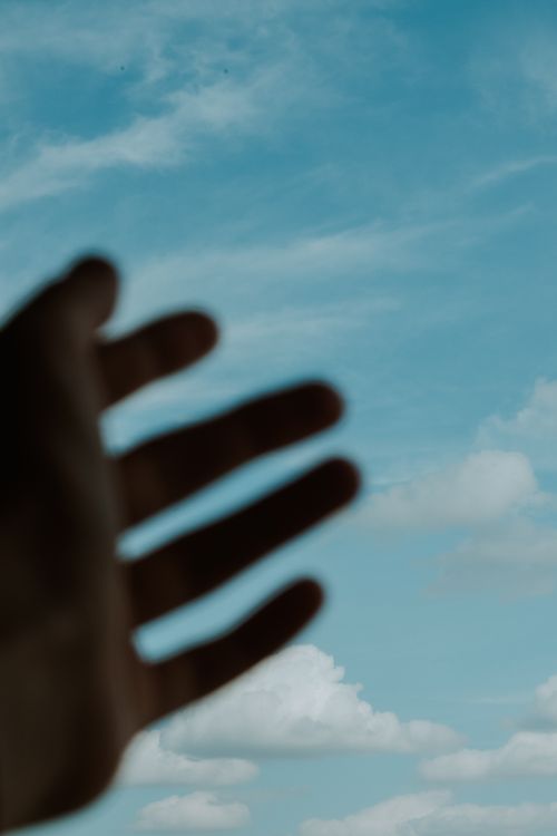 Free Blurred silhouette of a hand reaching towards a vibrant blue sky filled with fluffy white clouds. Stock Photo