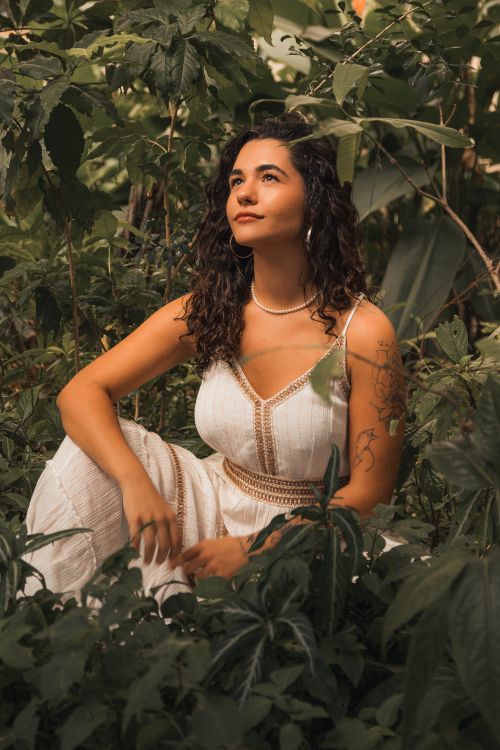 Free A woman in a white dress sits surrounded by lush green foliage, looking serene. Stock Photo