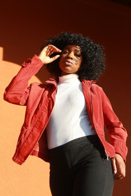 Stylish black woman in a red jacket posing outdoors with a confident look under natural sunlight.