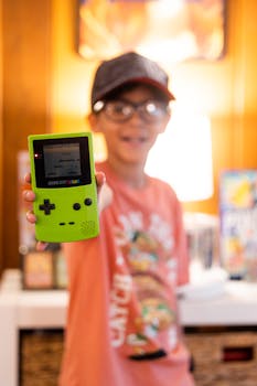 A young boy wearing a baseball cap and eyeglasses displays a vintage Game Boy Color handheld console indoors.