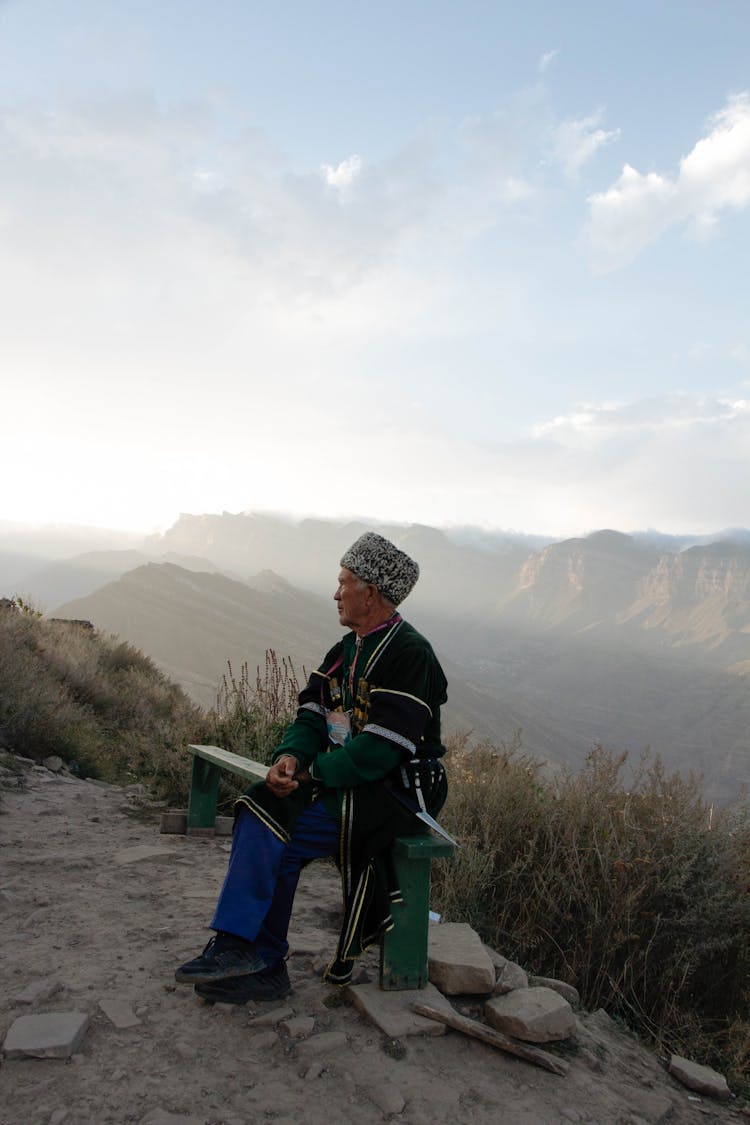 Senior Man Wearing Traditional Clothing Sitting In A Mountain Landscape