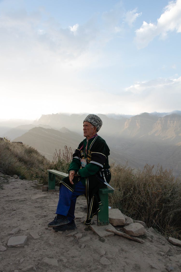 Senior Man Wearing Traditional Clothing Sitting In A Mountain Landscape