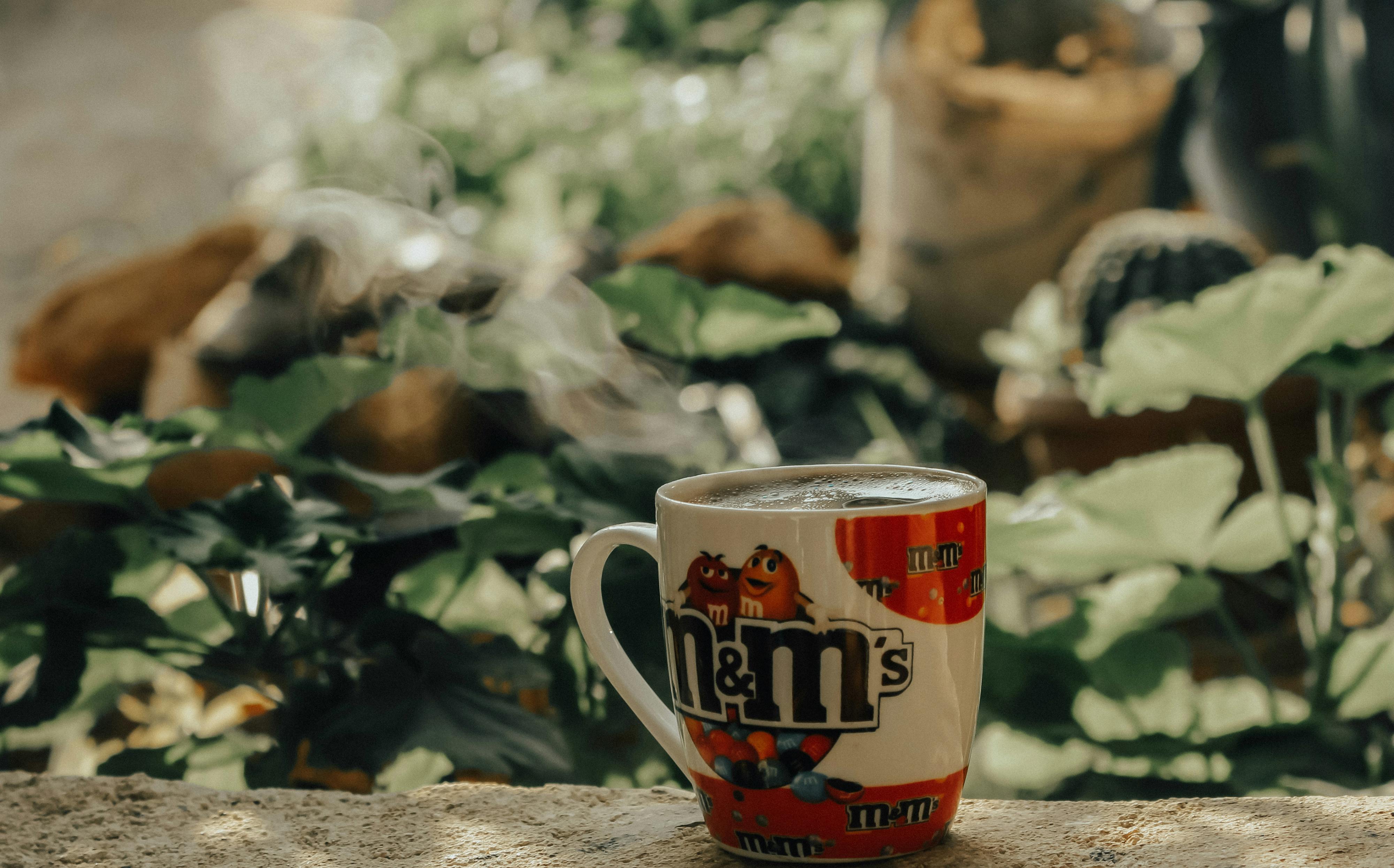 Hot coffee steaming in an M&M's mug amidst a serene outdoor garden setting.
