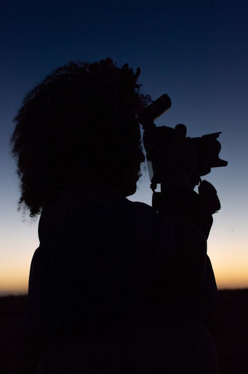 Silhouette of a Woman with Afro Hairstyle Photographing with a Camera at Sunrise