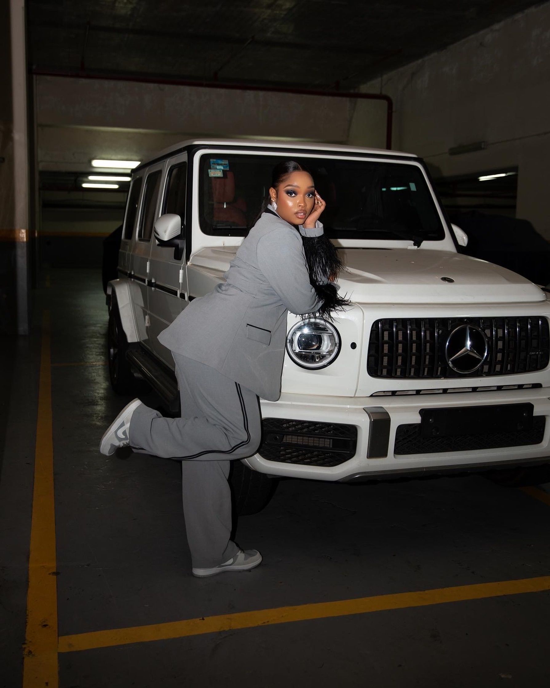 A fashionable woman in a gray suit poses confidently with a Mercedes G-Class SUV indoors.