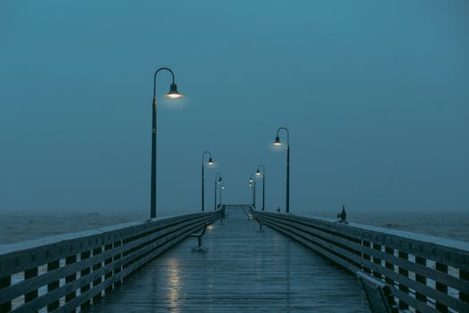 A scenic view of a rainy pier with glowing street lamps creating a moody atmosphere.