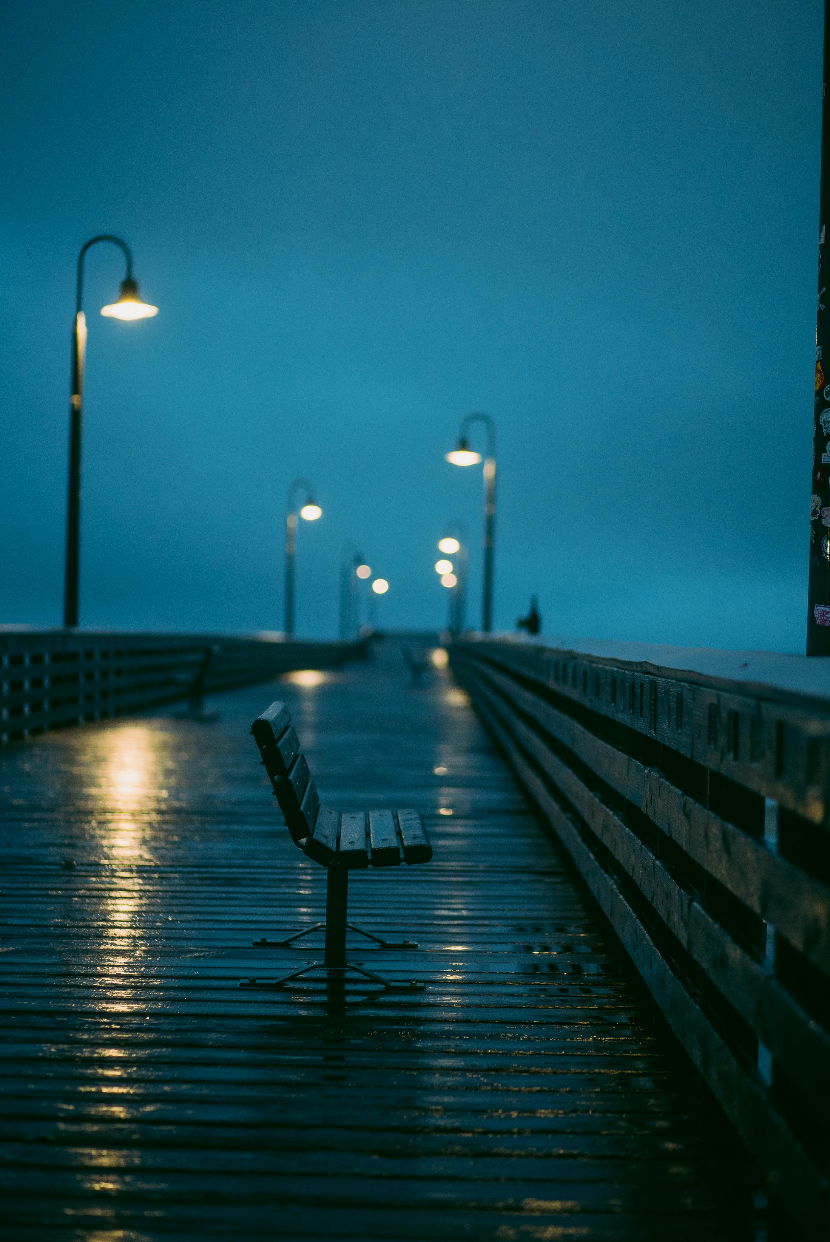 A quiet, rainy evening on an urban pier with street lamps illuminating the wet benches.
