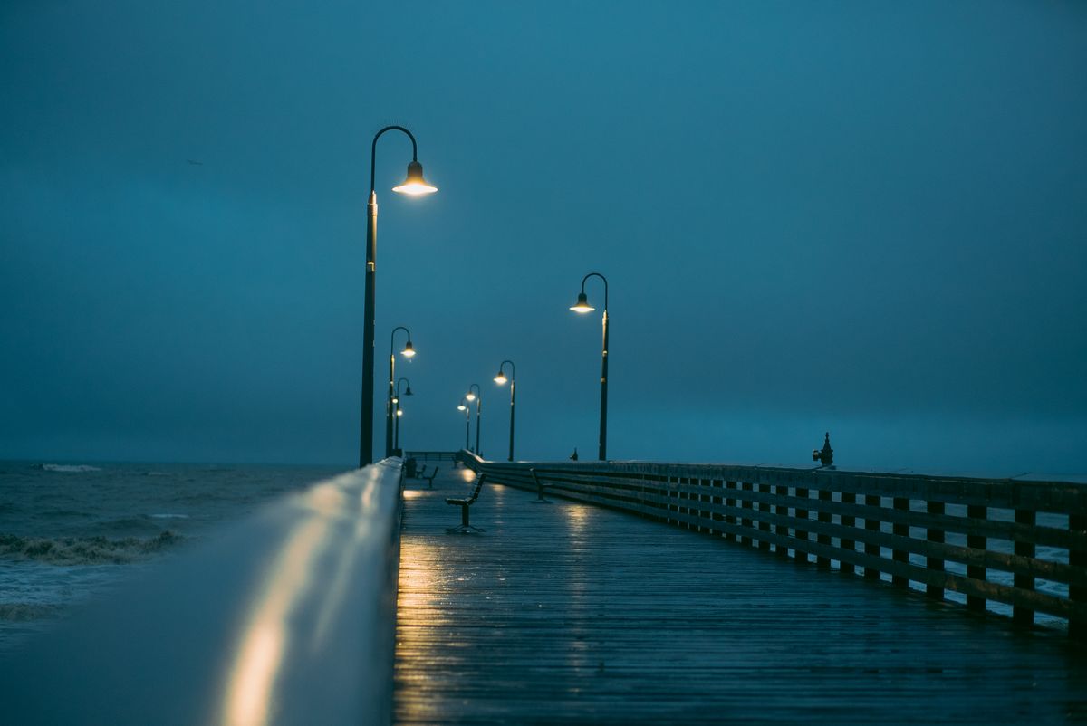 Wet seaside pier with glowing street lamps at dusk — photo by Josh Hild on Pexels