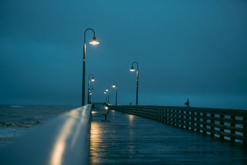 Free Dramatic view of a wet pier with glowing street lamps during an overcast evening by the sea. Stock Photo