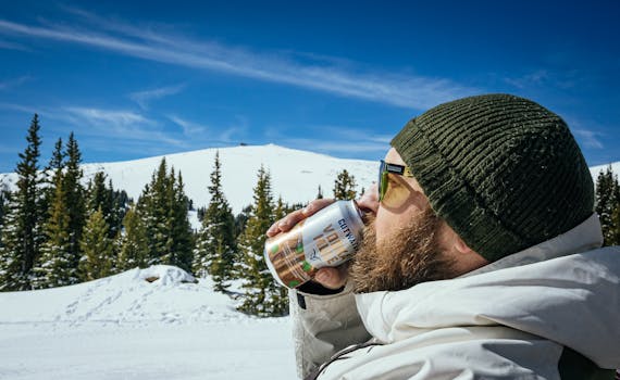 A man in a winter coat drinks from a can in a snowy mountain setting.