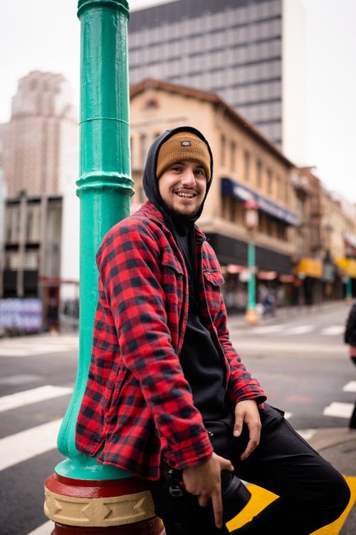 Free Smiling man in a red plaid jacket leaning on a colorful street pole in a city setting. Stock Photo