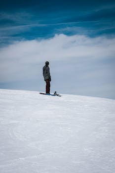 A lone snowboarder stands on a snowy slope under a bright blue sky, capturing the essence of winter adventure.