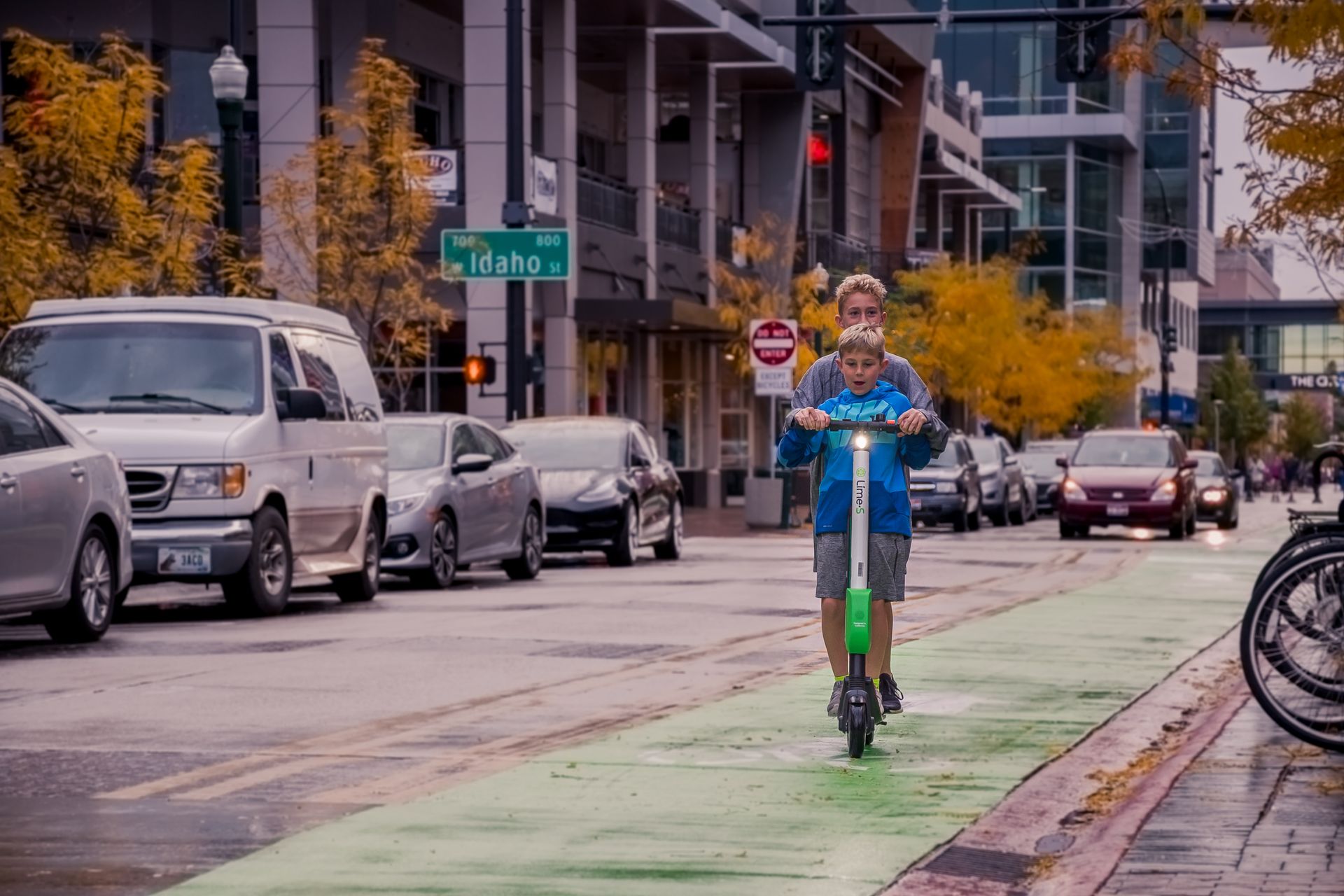 Two boys riding an electric scooter on a city street with autumn trees and traffic.