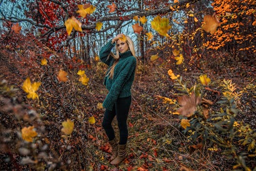 A beautiful blonde woman in a thick sweater stands amidst falling autumn leaves in a forest.