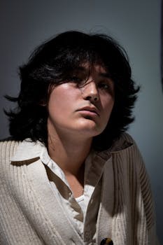 Moody portrait of a young man with long black hair in a cream sweater indoors.