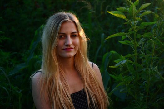 A beautiful blonde woman smiling in a natural outdoor setting with lush greenery.