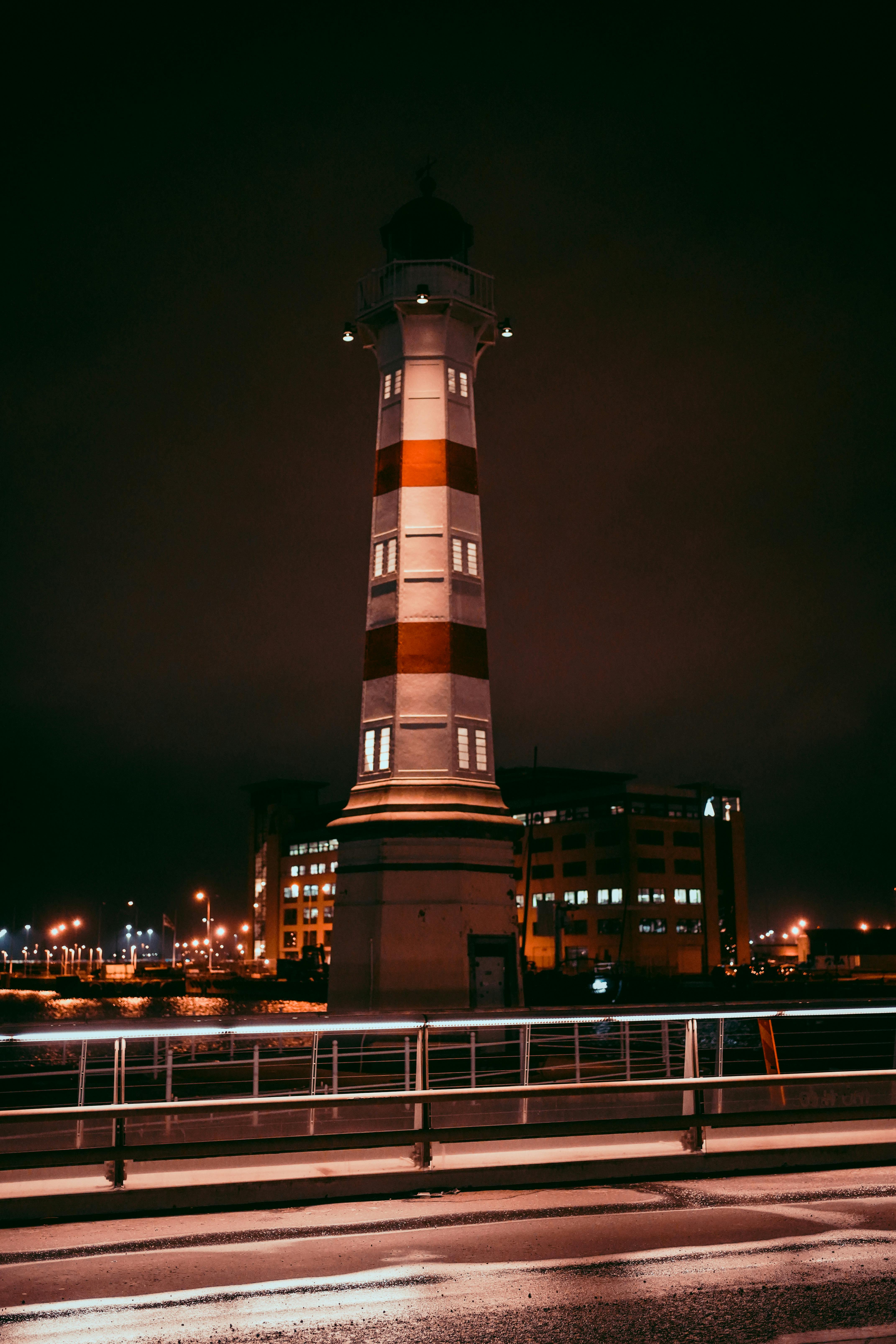 A striking lighthouse illuminated during the night in a cityscape, providing navigation aid.