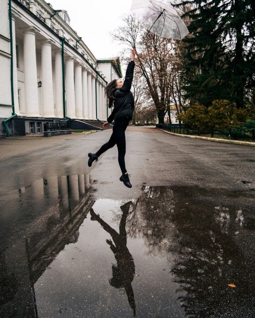 Free A woman leaps joyfully with an umbrella on a rainy street, capturing her reflection in a puddle. Stock Photo