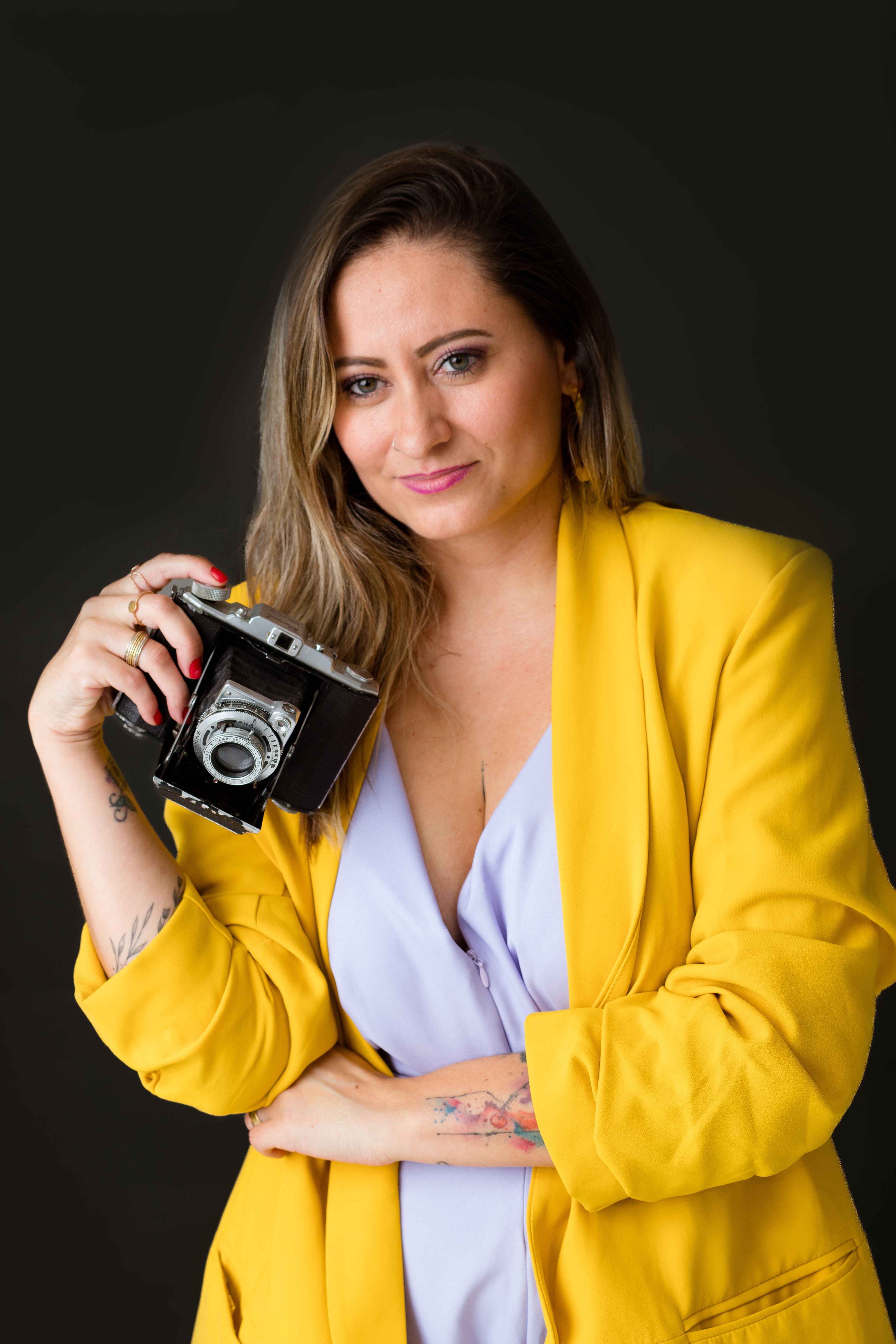 Stylish woman posing with a vintage camera against a dark background wearing a yellow blazer.