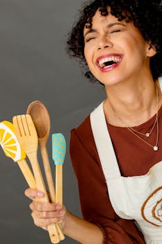 Laughing woman with curly hair holding vibrant kitchen utensils against gray backdrop.