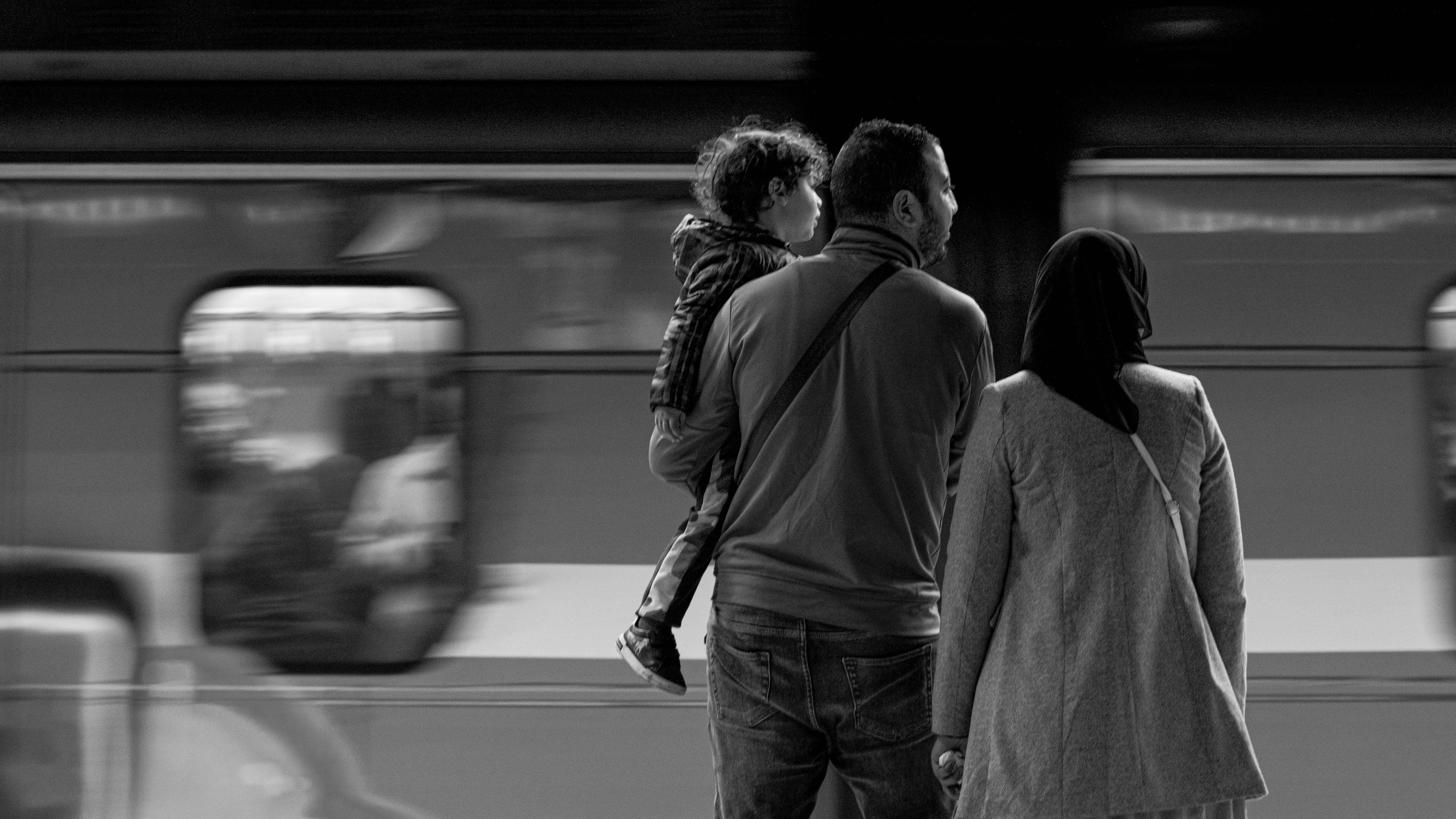 A family waits at an urban subway station with a train passing by in blurred motion.