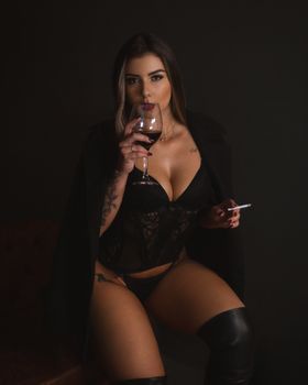 Seductive model in lingerie enjoying wine and cigarette in studio setting.