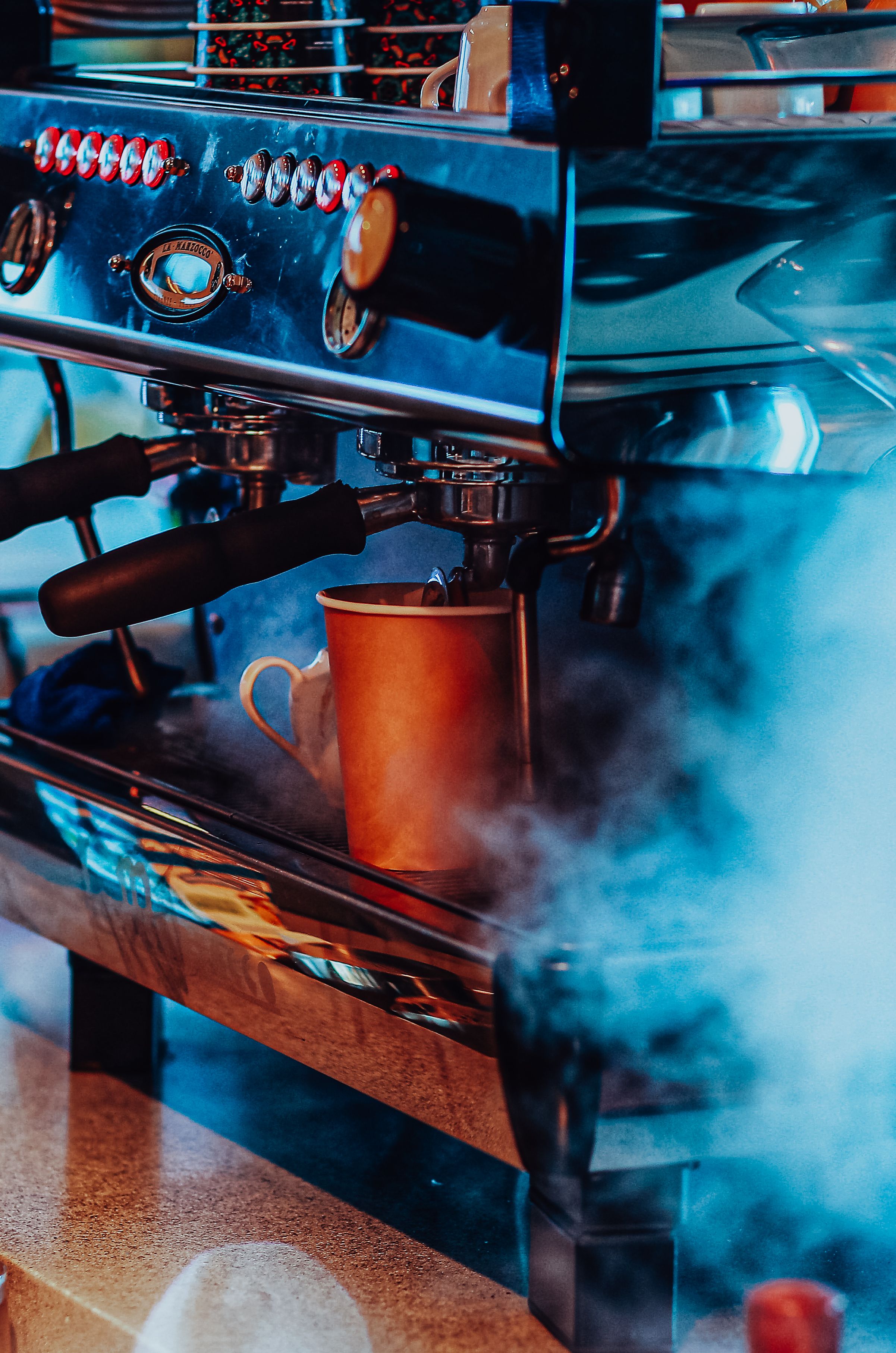 Close-up of a steaming mug under an espresso machine in a vibrant café setting.