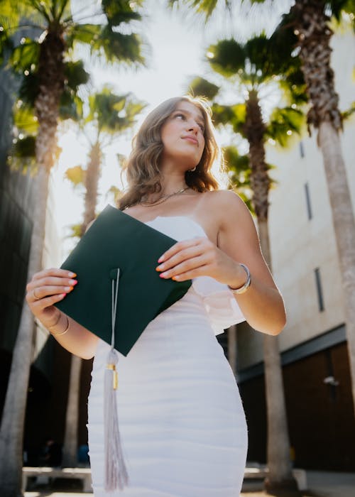 Young Woman in a White Dress Holding a Mortarboard and Standing near Palm Trees
