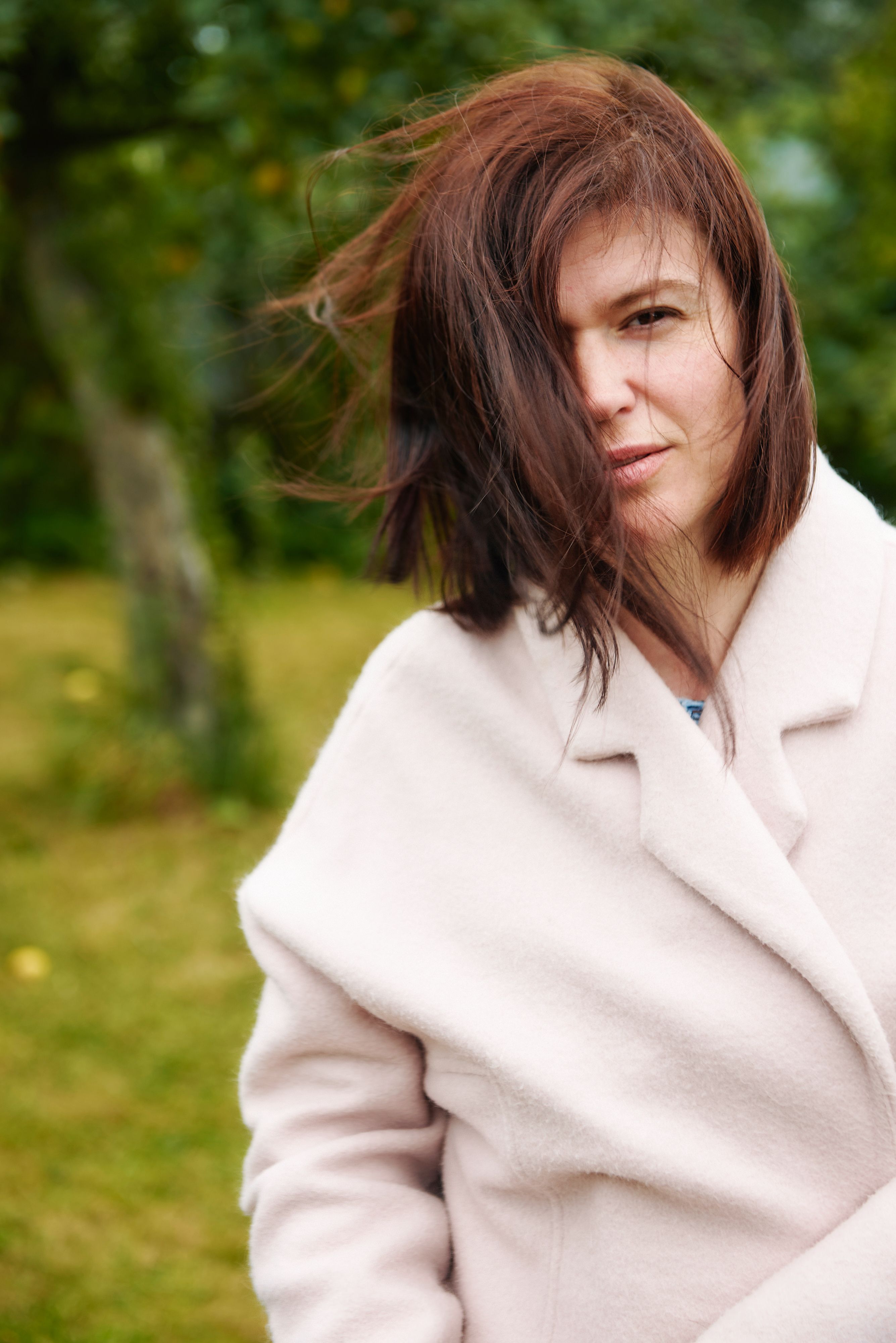 Portrait of a woman with brown hair wearing a coat in an outdoor park setting.