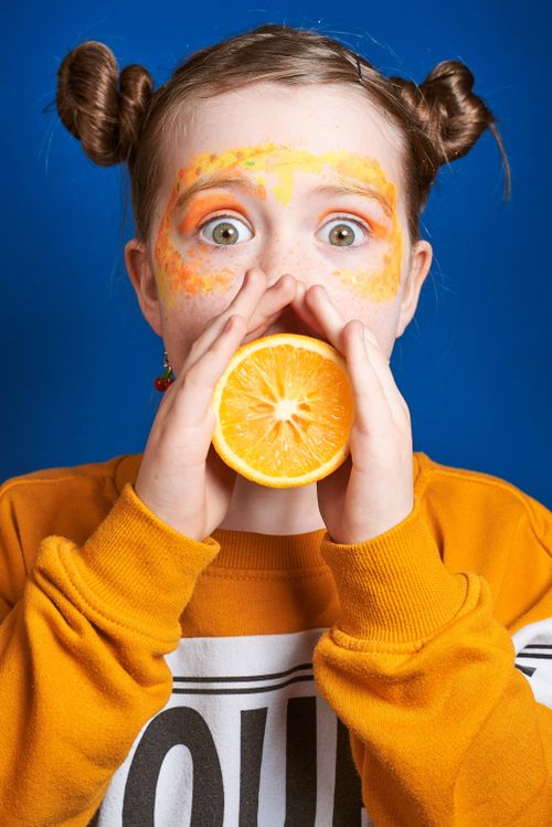 Free A playful child in a colorful sweater holding a halved orange against a vivid blue background. Stock Photo