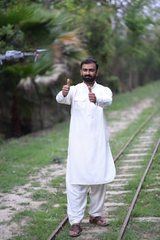 Adult man in traditional attire giving thumbs up next to a drone on grassy tracks.