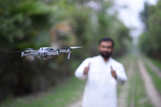 A close-up of a drone flying outdoors with a man gesturing thumbs up in the background.