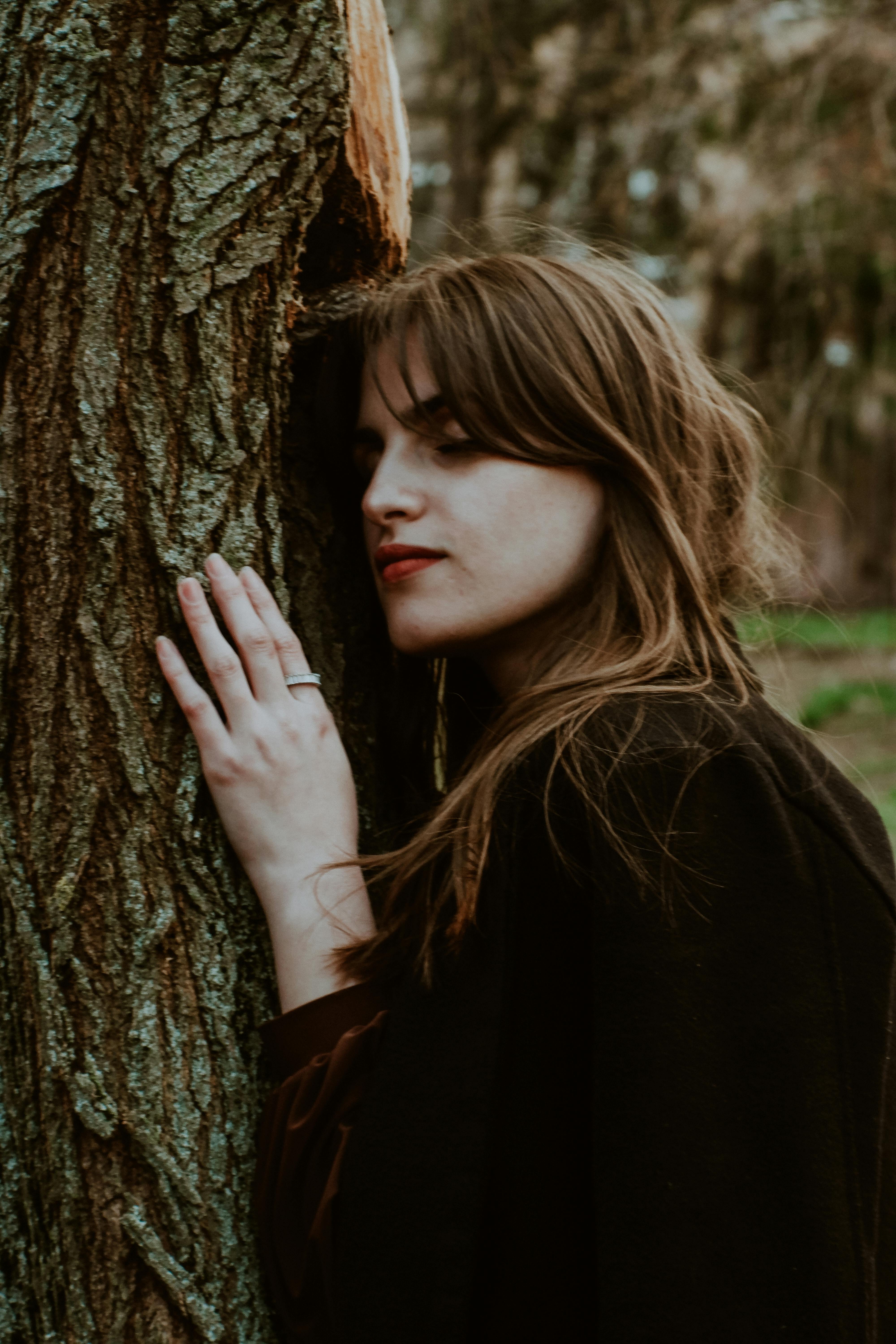 A serene woman connects with nature, gently touching a tree trunk, embodying tranquility and harmony.