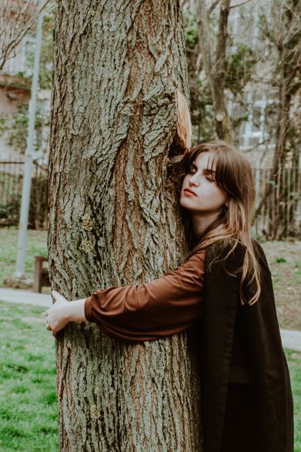 A woman with brown hair gently embraces a tree in a tranquil park environment.