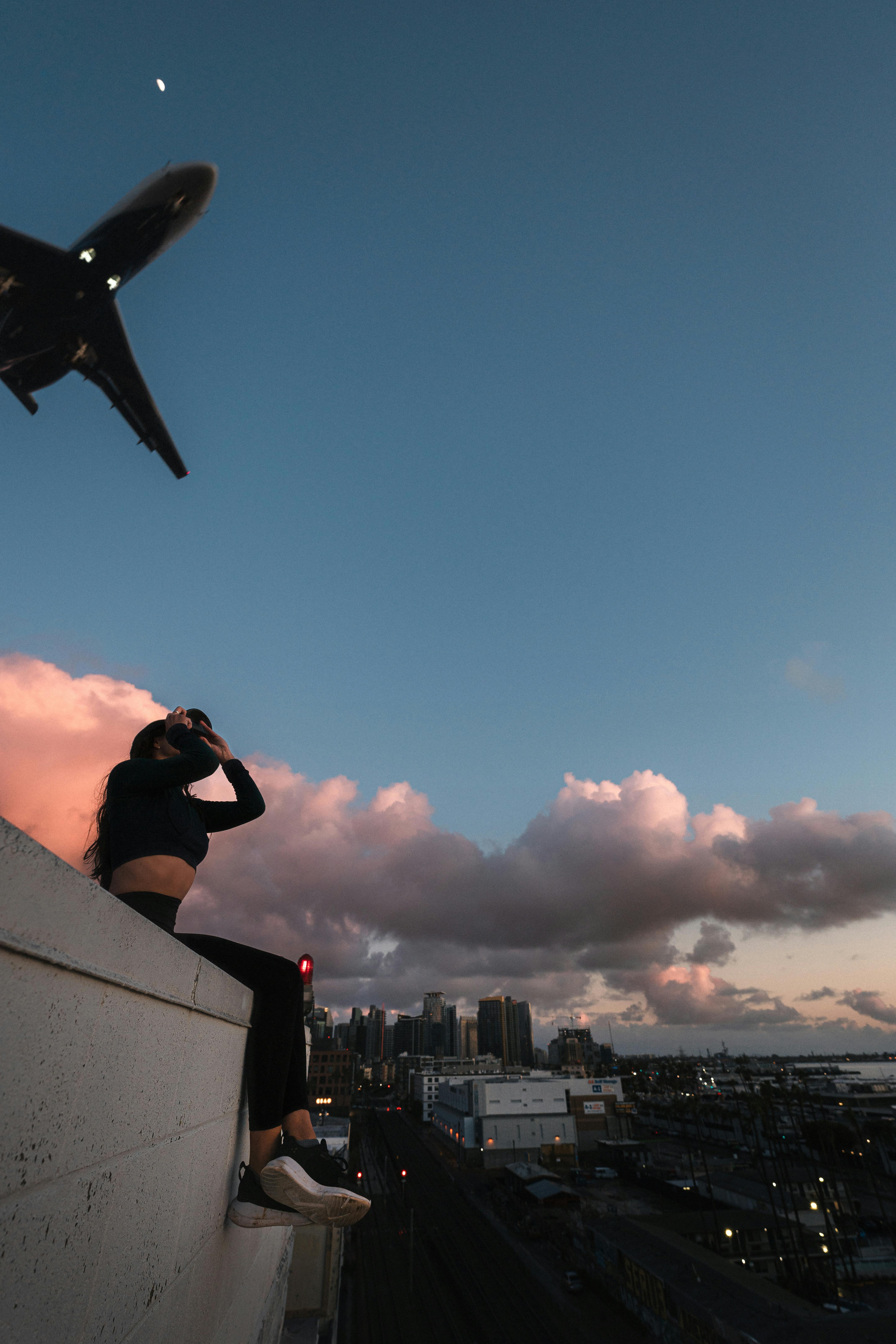 A woman sits on a rooftop in San Diego at sunset, watching an airplane pass by.
