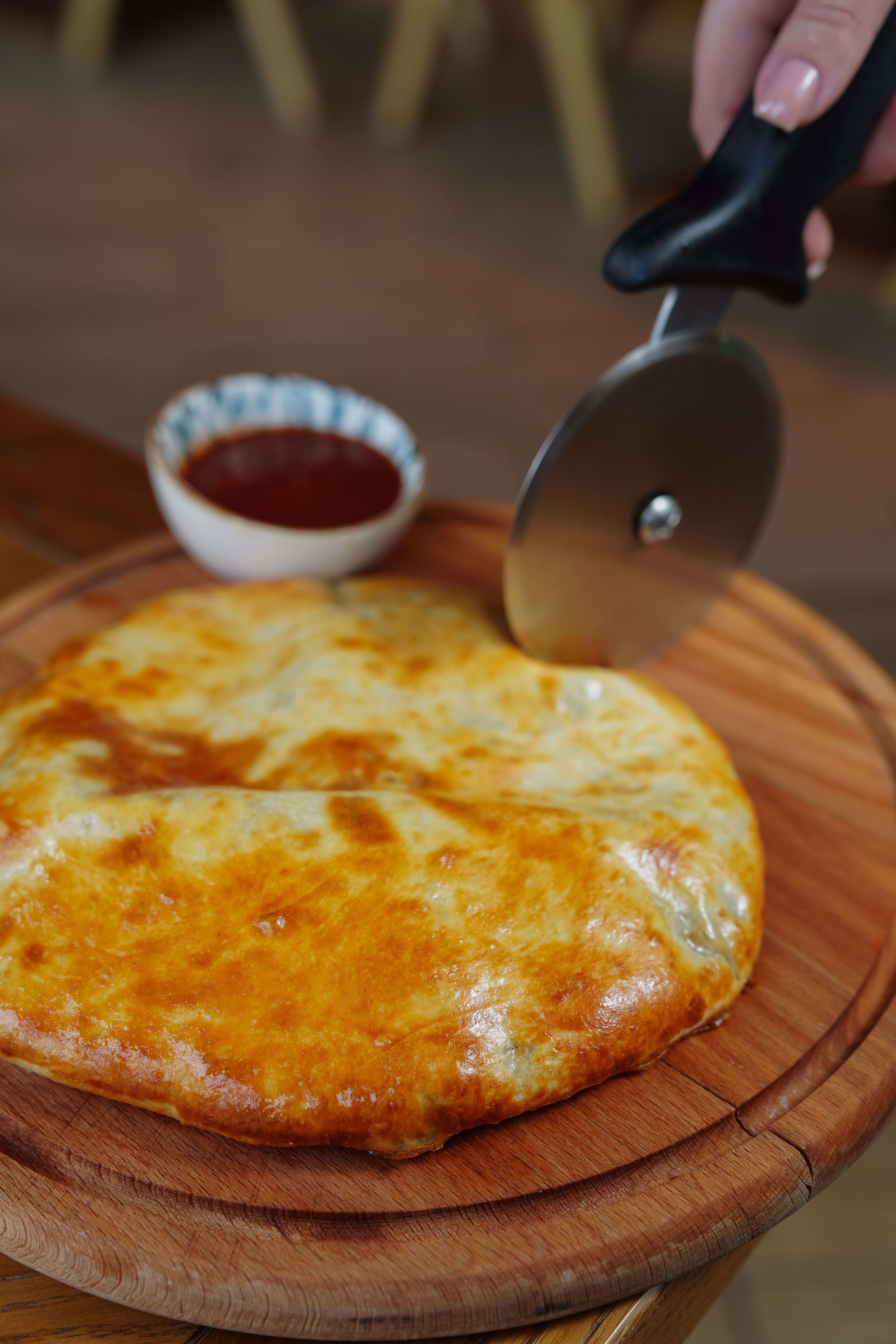 Khachapuri on a wooden board being sliced with a pizza cutter, accompanied by dipping sauce.