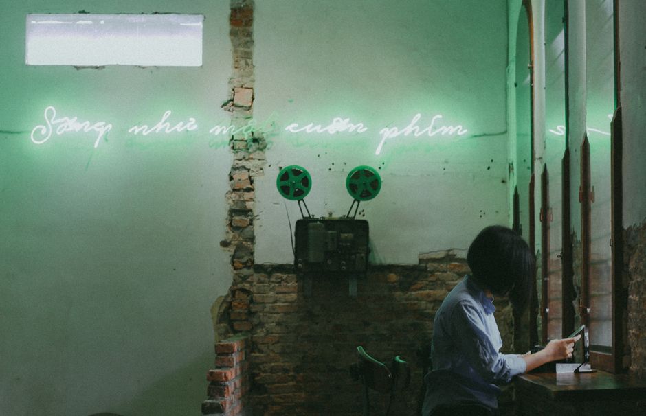 A woman checks her cellphone in a vintage room with neon signs and exposed brick walls.
