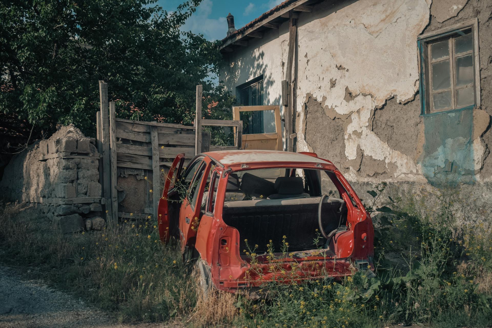 An abandoned red car sits by a rustic rural house with damaged walls under clear skies.