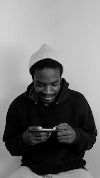 Black and white portrait of a young man smiling while using his smartphone, wearing a hoodie and beanie.