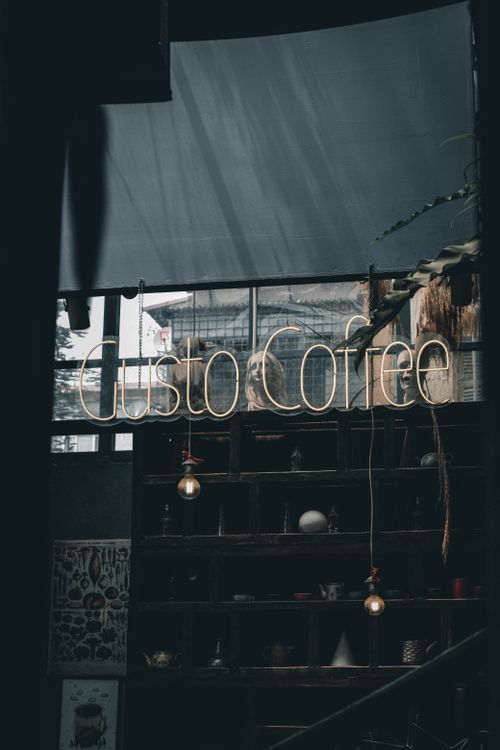 Free Moody urban café interior featuring a neon 'Gusto Coffee' sign and rustic shelves. Stock Photo