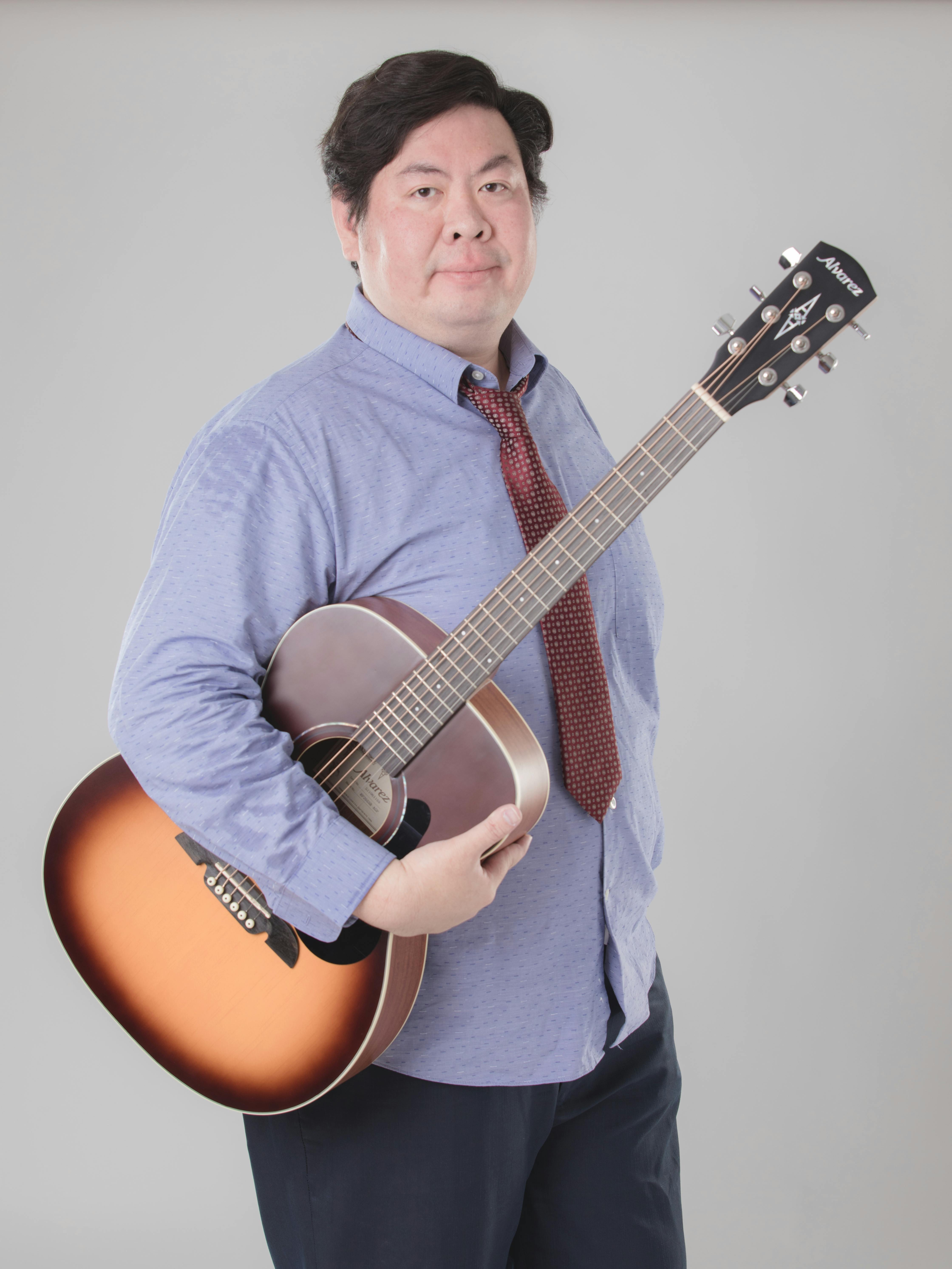 Asian man with guitar in casual attire posing in photo studio. Ideal for fashion and music themes.