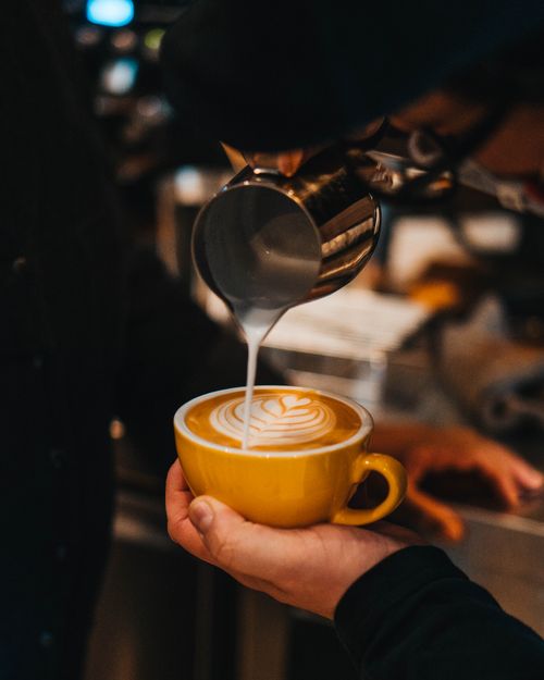 Free Close-up of a barista pouring milk to create heart latte art in a yellow cup at a cozy café. Stock Photo