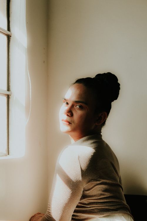 Free A woman gazes thoughtfully as warm sunlight filters through the window. Stock Photo