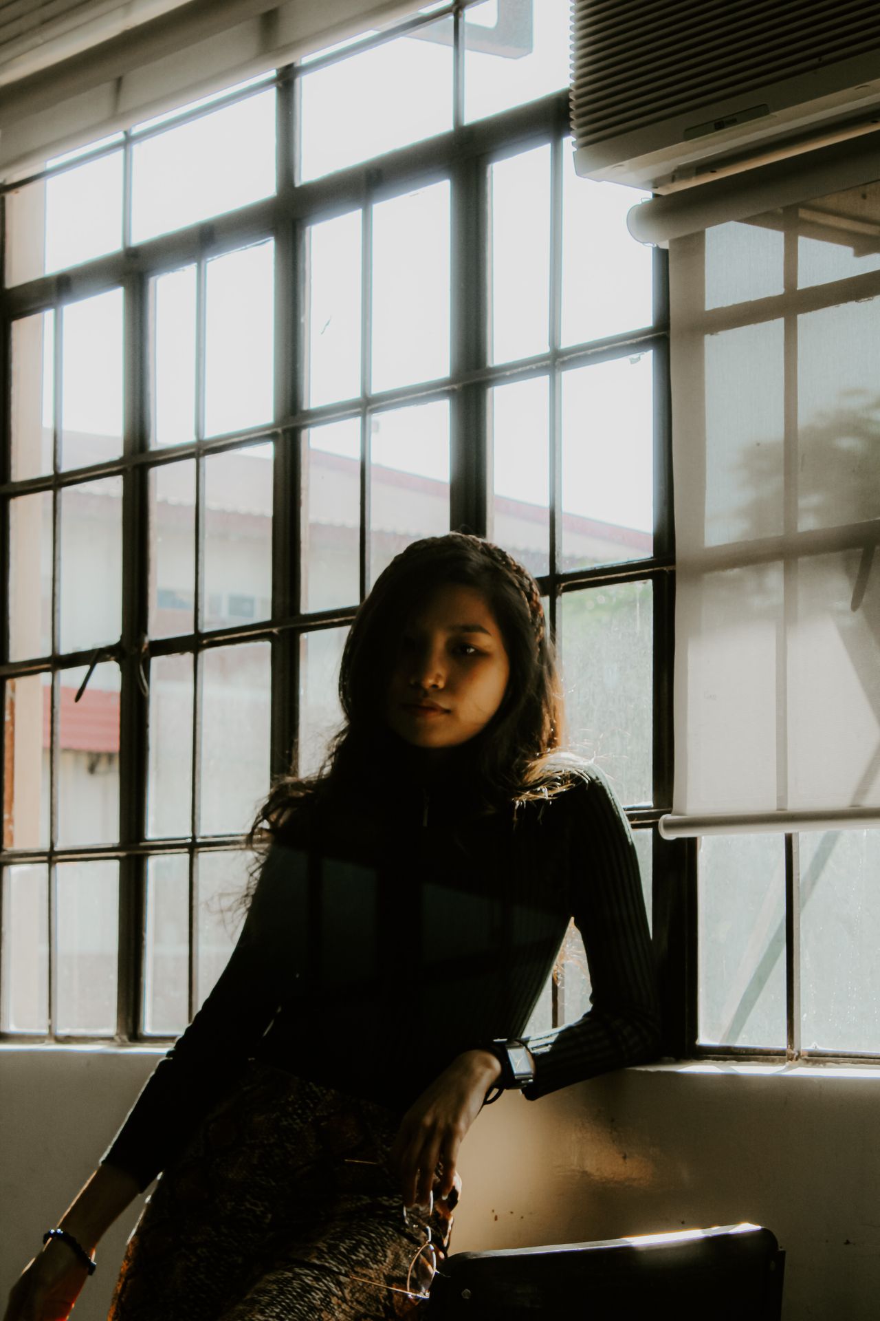 A young woman poses inside with dramatic lighting through large windows